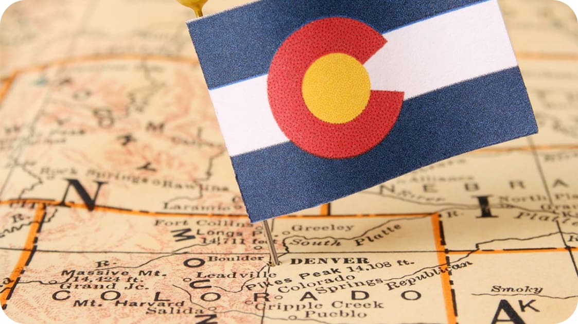 A small Colorado flag is stick-pinned into a vintage map, marking the state. The map displays various cities and geographical details of Colorado and neighboring areas.