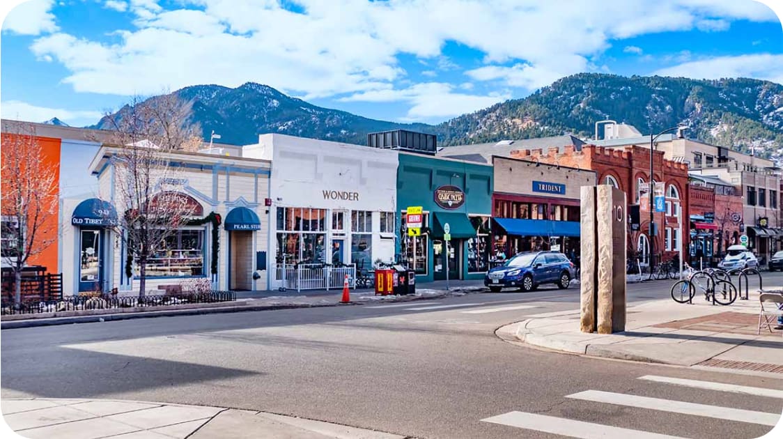 A street scene in a small town with colorful storefronts, including a bakery and a restaurant. Snow-capped mountains and a clear blue sky can be seen in the background. A pedestrian crossing is visible in the foreground.